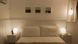 Een net opgemaakt bed met witte lakens en twee bedlampjes. Boven het bed hangen drie kleine vierkante spiegels aan de muur.