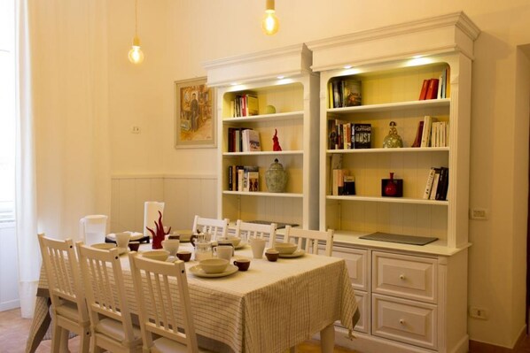 Un coin repas avec une table dressée pour quatre personnes, entourée de chaises blanches. On y trouve deux étagères blanches, ornées de livres et d’objets décoratifs.