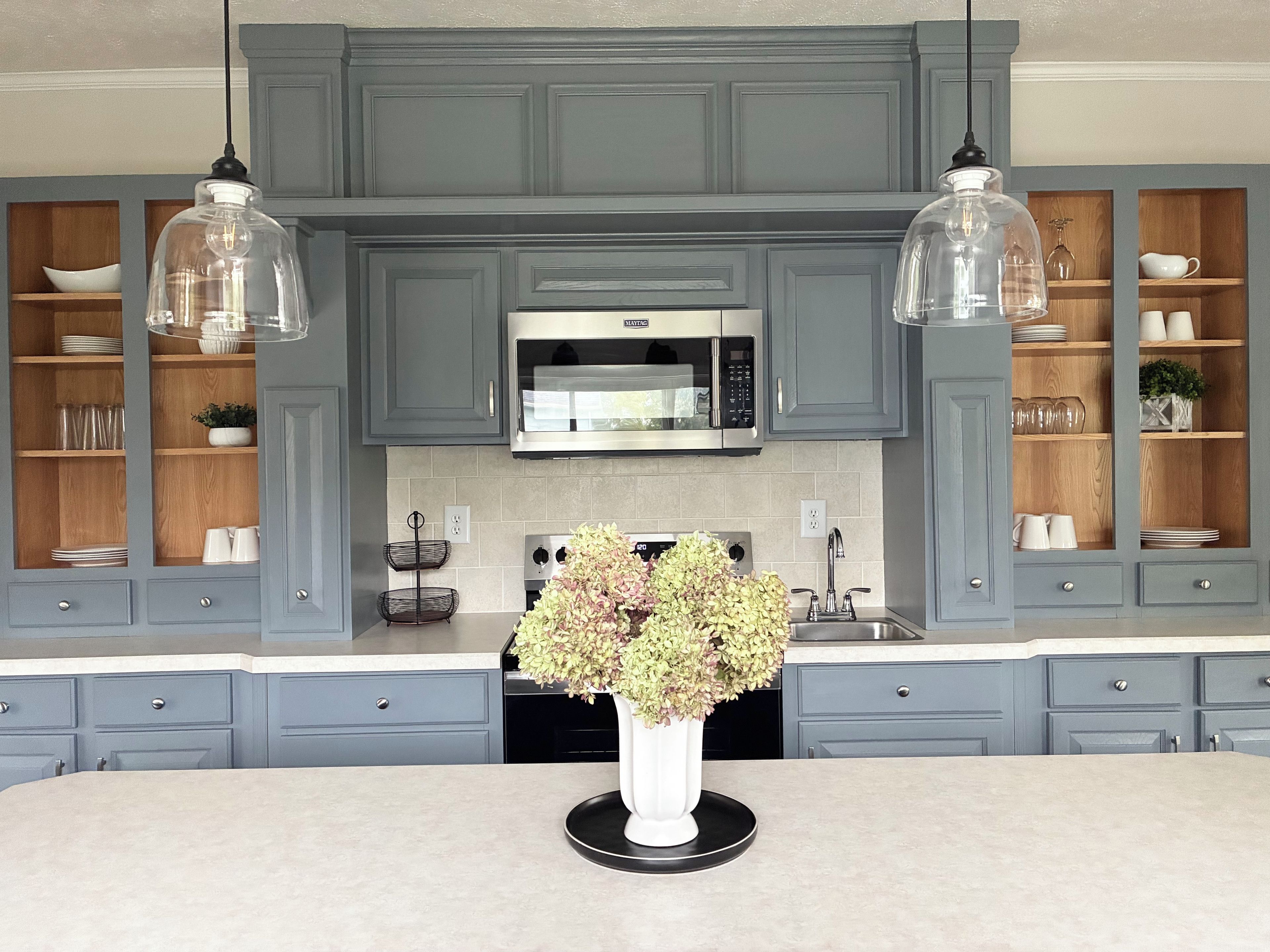 A kitchen with grey cabinets, a central island with a vase of flowers, and pendant lights.
