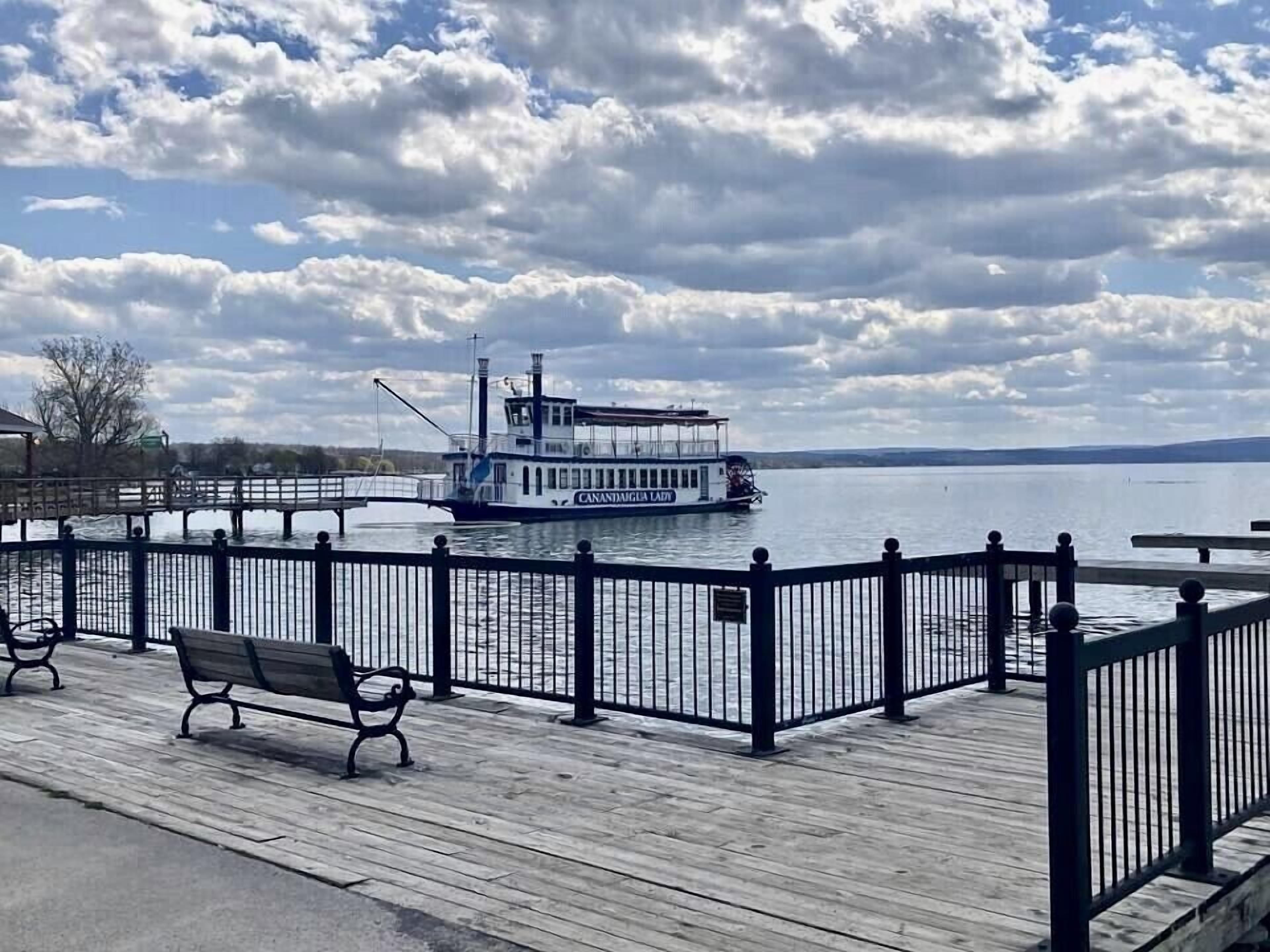 Een houten steiger met bankjes, een boot genaamd "CANANDALE LA LADY" op het water, en een bewolkte lucht.