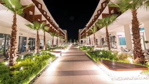 A well-lit pathway flanked by palm trees and greenery, leading to a building with a modern design and multiple floors. - Liberty Fabay - All Inclusive  (Fethiye)