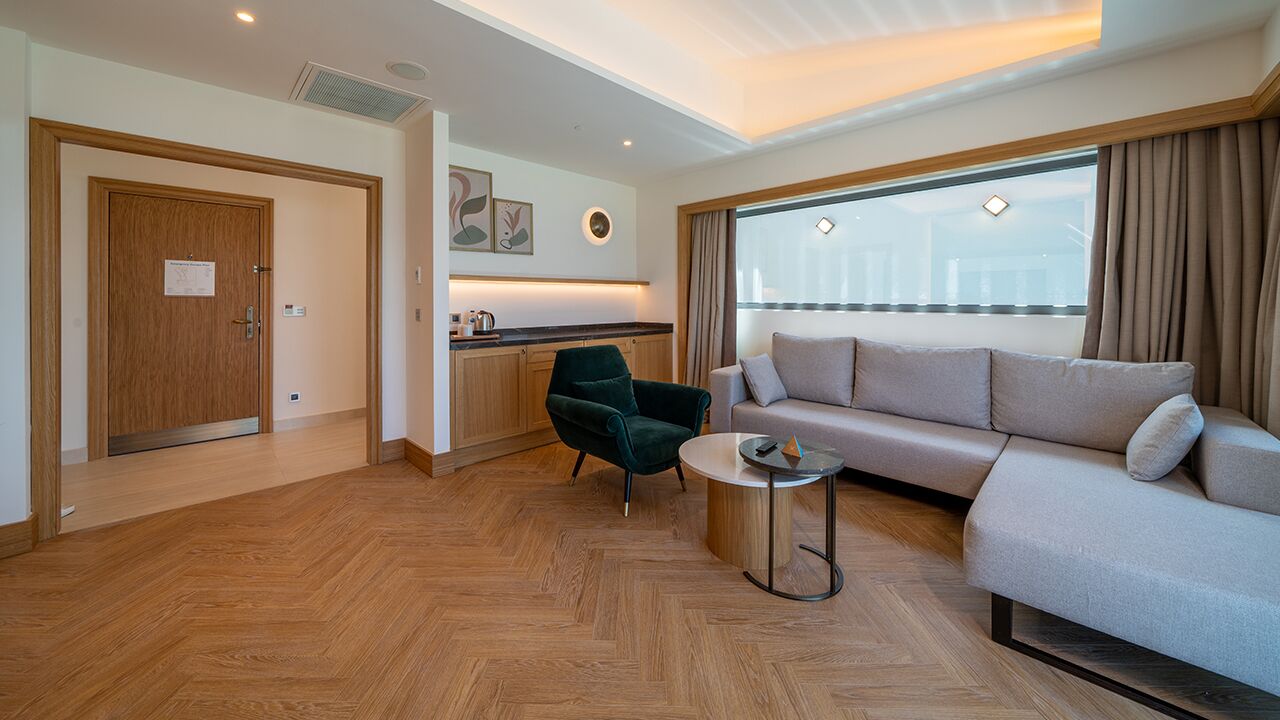 A modern hotel room with a wooden floor, a sofa, a green armchair, a small round table, and a large window with curtains.