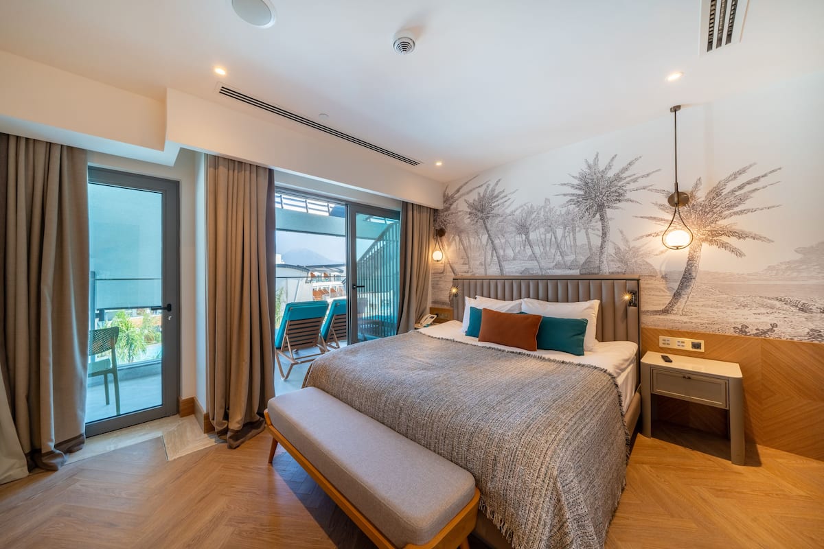Une chambre d’hôtel moderne avec un grand lit, une banquette, une table de chevet et une fresque représentant des palmiers.