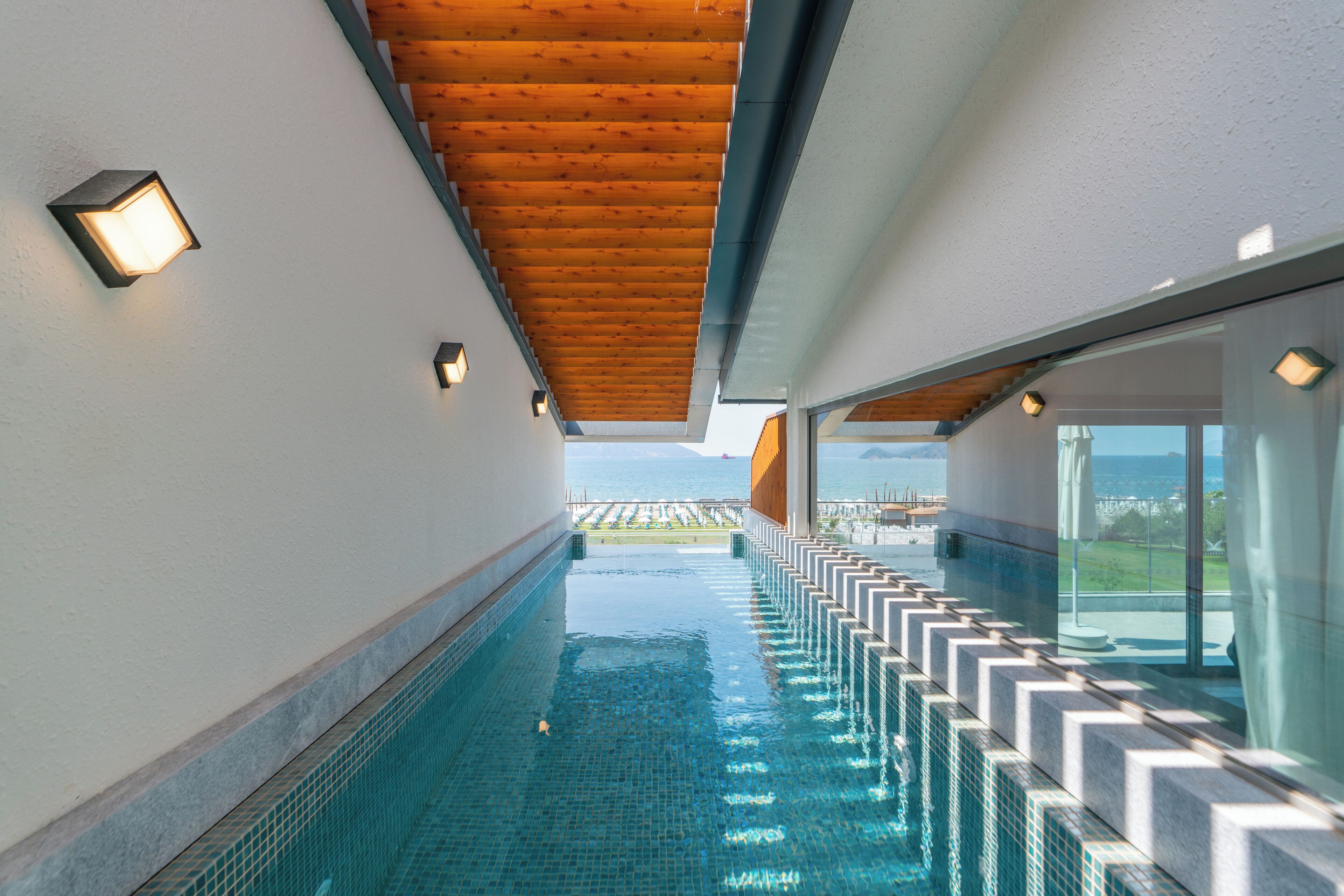 A modern indoor swimming pool with a wooden ceiling and glass walls offering a view of the sea.