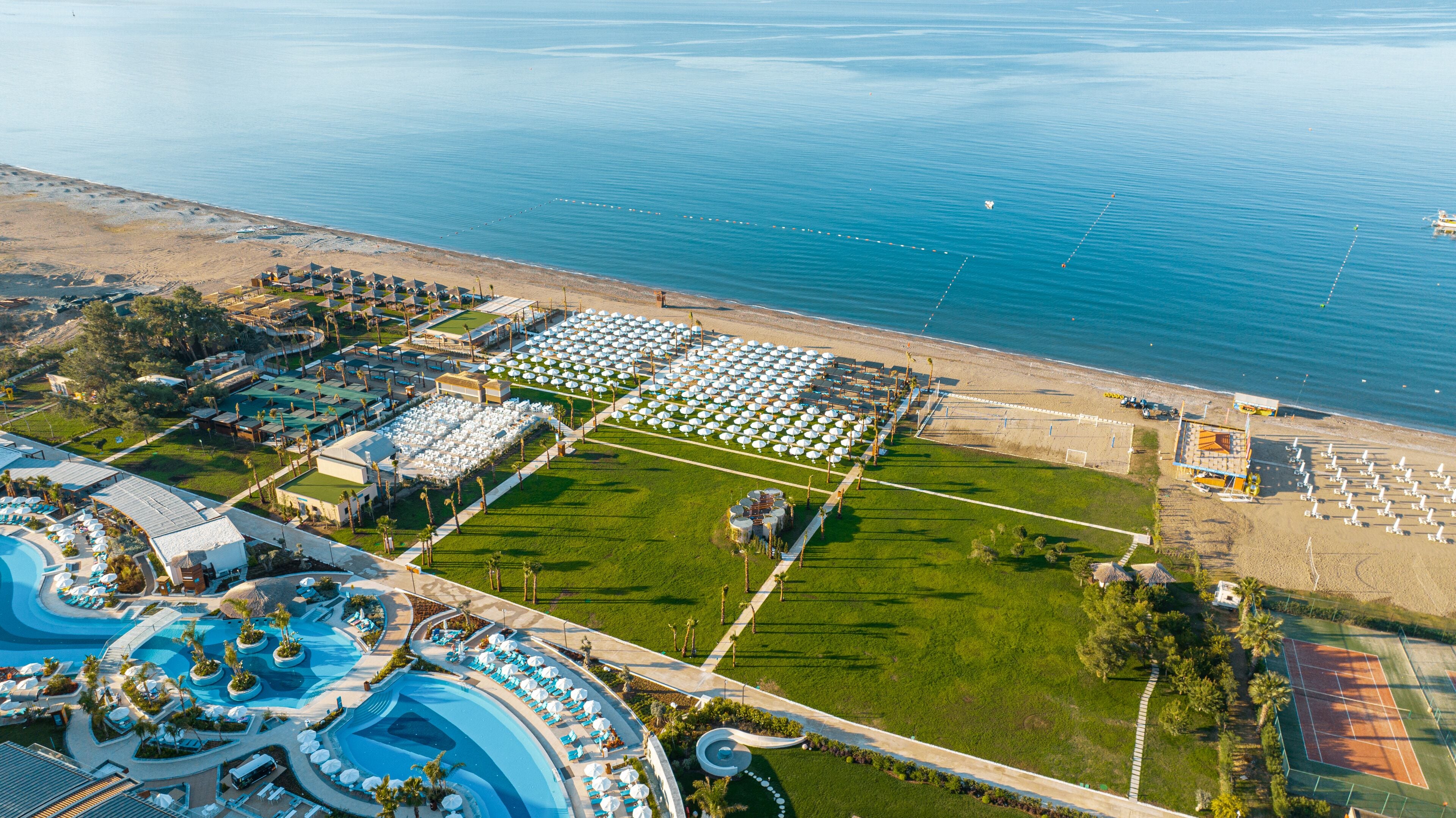 An aerial view of a resort with a large pool area, multiple tennis courts, and a beach with umbrellas.