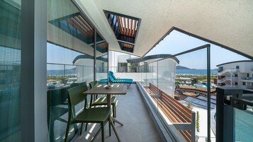 A balcony with a table and chairs, a blue lounge chair, and a view of the sea and buildings.