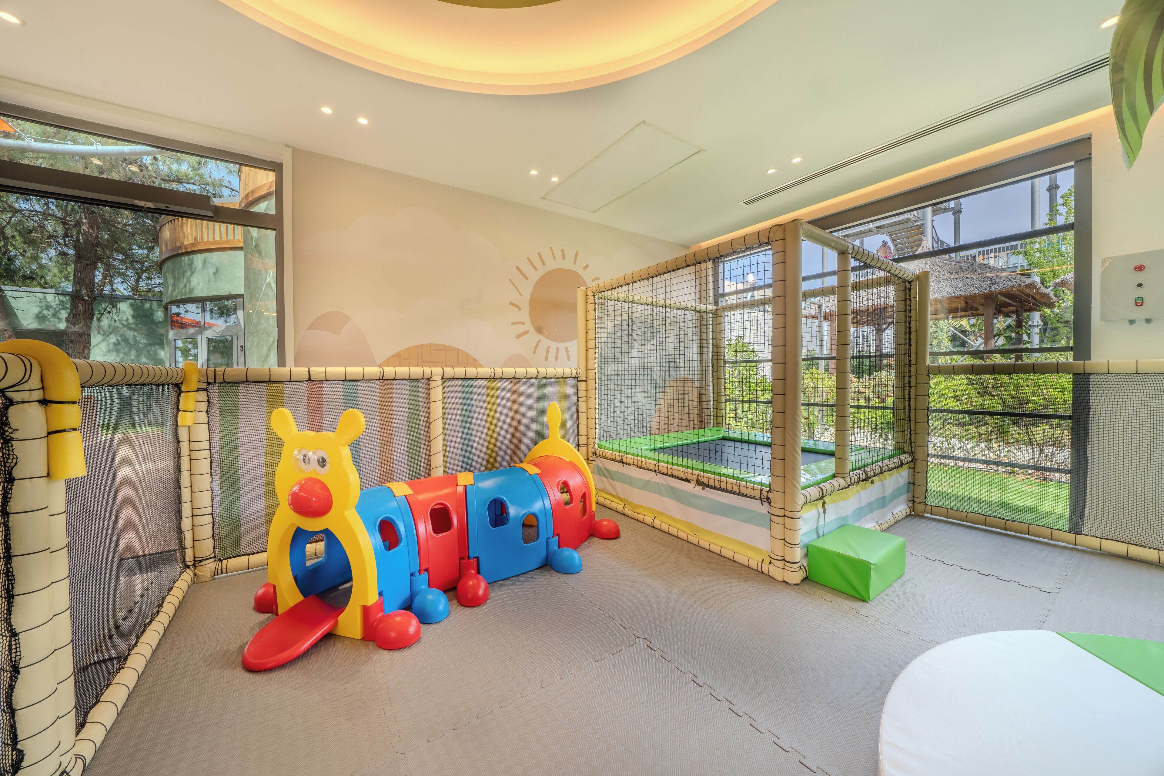 A play area with a colorful train set, padded flooring, and a view of the outdoors through a large window.