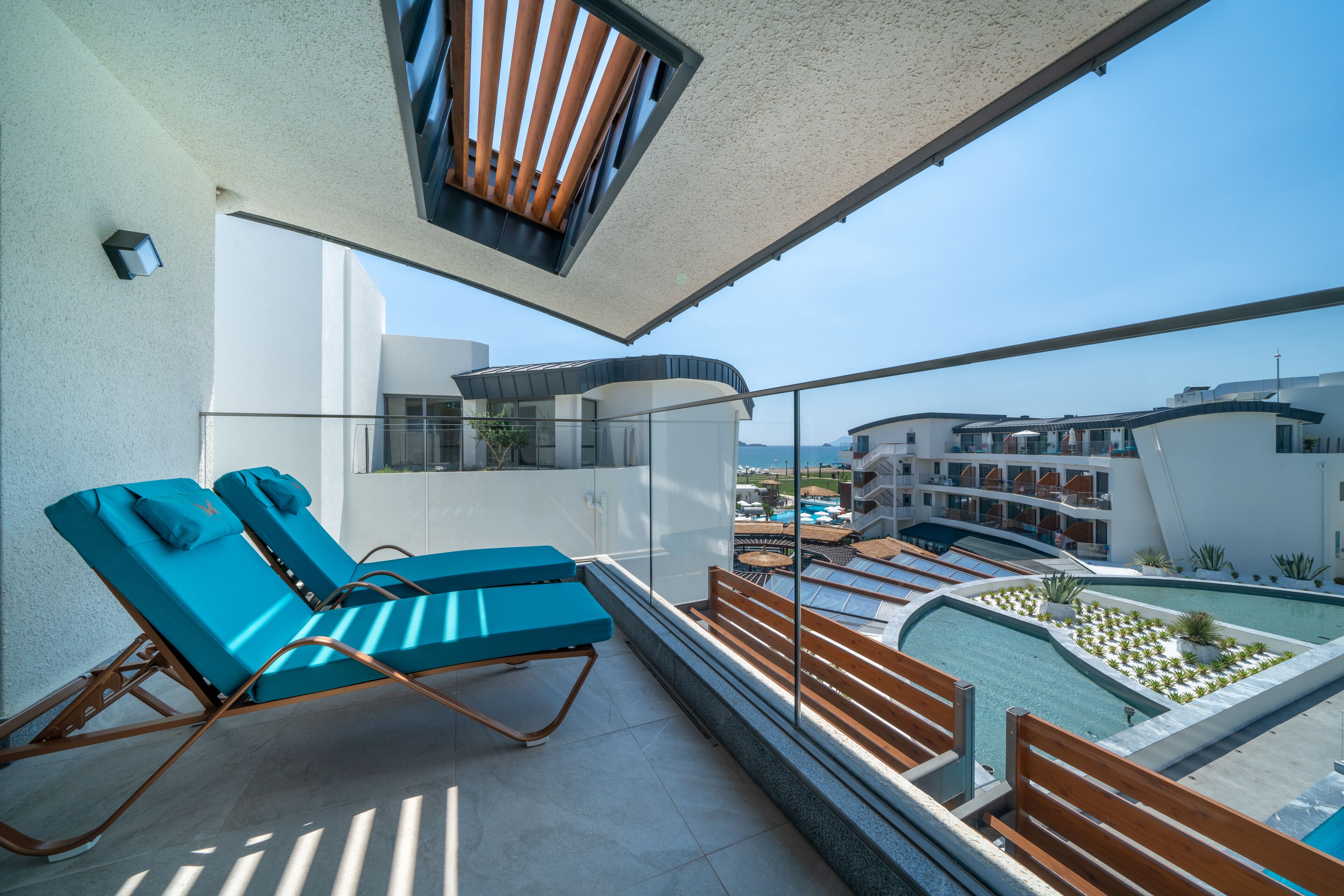 A balcony with two blue lounge chairs, a glass railing, and a view of a pool and buildings.