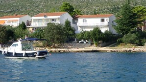 Un hôtel situé sur la côte, composé de plusieurs bâtiments blancs, avec un bateau amarré à proximité et une colline rocheuse en arrière-plan.