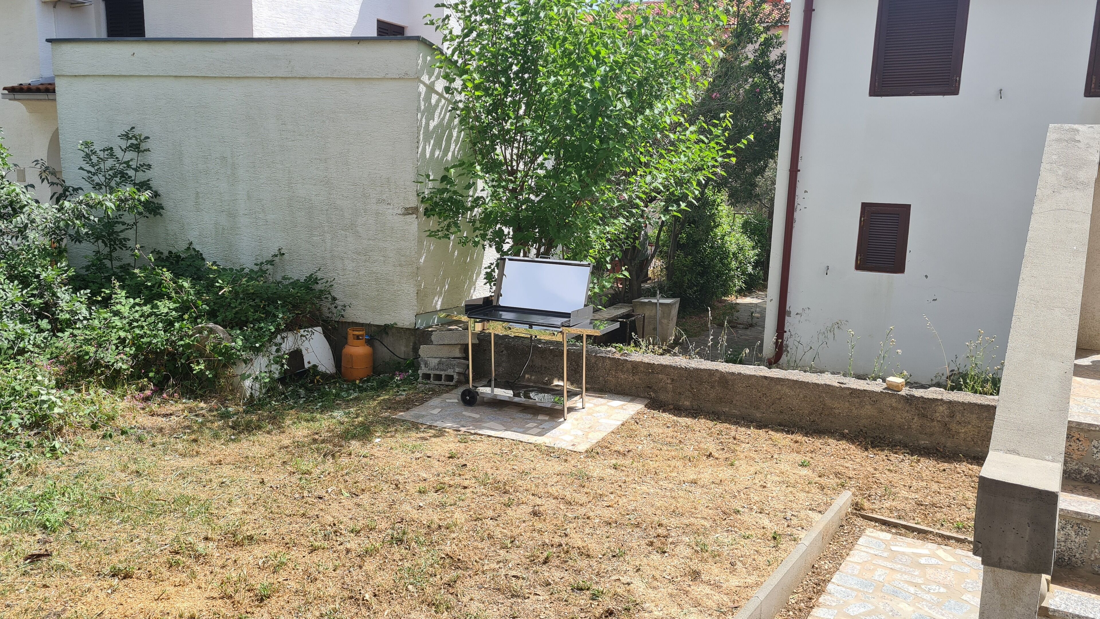Ein Garten mit einem kleinen Tisch, einem Gasflaschen und viel Grün, umgeben von einem weißen Gebäude.