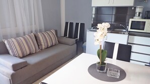 Un salon moderne avec un canapé gris, des coussins à rayures, une table à manger agrémentée d’un vase de fleurs blanches et un coin cuisine équipé d’appareils électroménagers.