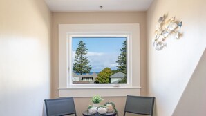 Habitación de esquina con ventana que da a árboles y un cielo despejado, dos sillas, una mesita con adornos y una lámpara fijada a la pared.