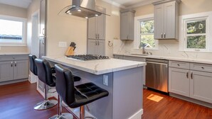 Una cocina moderna con armarios grises, una isla central con taburetes de bar, electrodomésticos de acero inoxidable y un ventanal que proporciona luz natural.