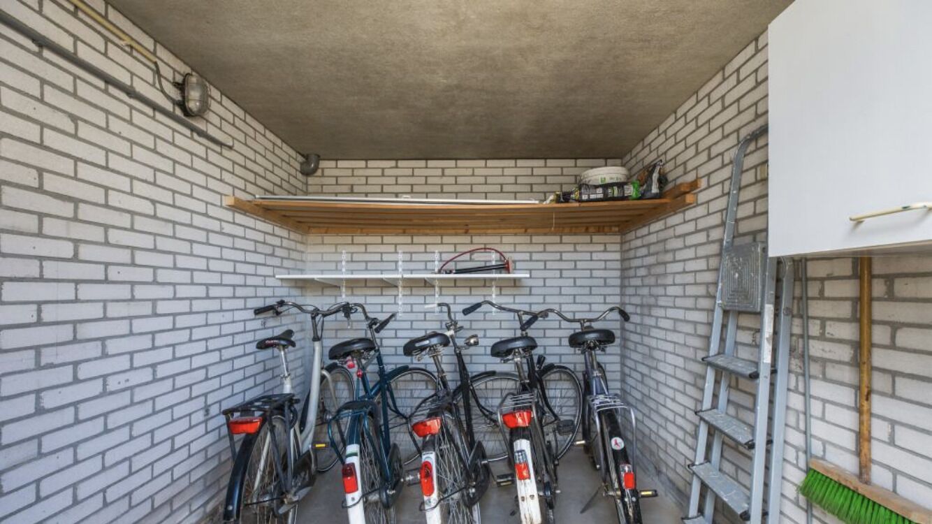 A narrow, enclosed space with a brick wall and a wooden shelf. Several bicycles are parked against the wall, and a ladder is leaning against the wall.