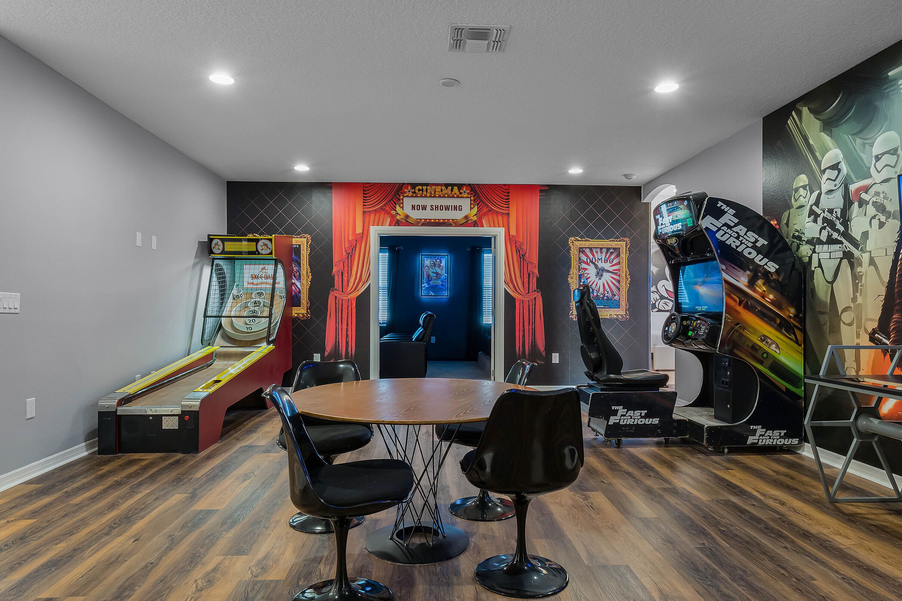 Una habitación con piso de madera, una mesa redonda con cuatro sillas negras, una zona de juegos con máquinas de arcade y una puerta que conduce a otra habitación.