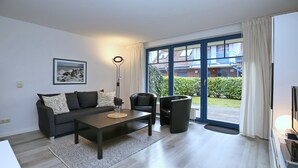 Ein modernes Wohnzimmer mit einer Couch, einem Couchtisch und Sesseln, einem großen Fenster mit Vorhängen und einem gerahmten Bild an der Wand.
