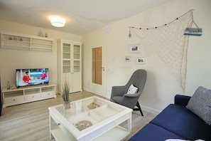 Ein Wohnzimmer mit Fernseher, einem Glastisch, einem blauen Sofa, einem grauen Sessel und einem weißen Schrank.