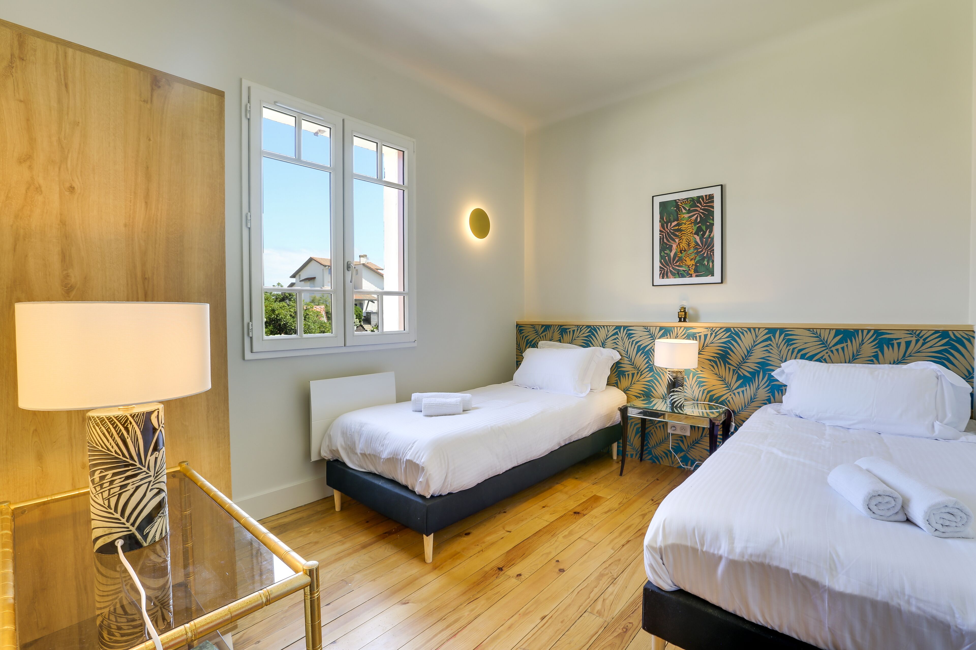 A hotel room with two beds, a window, a wooden wall, a framed picture, and a bedside table with a lamp.