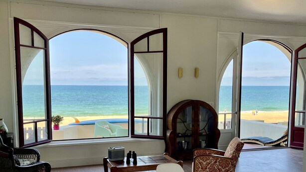 Una habitación con amplios ventanales de arco que dan a una playa, un armario de madera y una silla de descanso.