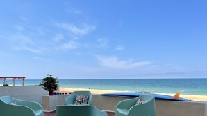 Un patio frente a la playa con sillas verdes y una tabla de surf, con vista a una playa de arena y un mar tranquilo.