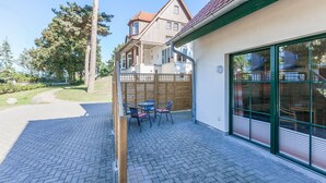 Eine Terrasse mit Backsteinpflaster, Holzlattenzaun und Blick auf ein traditionelles Haus im Hintergrund.