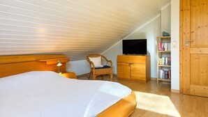 Un dormitorio con techo inclinado, una cama, una silla, un aparador, un televisor y una estantería.