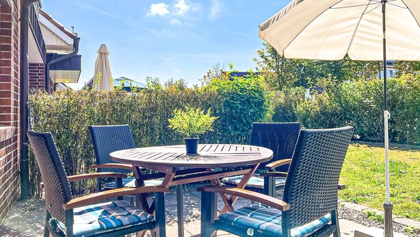 Eine Terrasse mit einem runden Tisch und Stühlen, einem großen Sonnenschirm und Blick auf ein Gebäude sowie Grünflächen im Hintergrund.