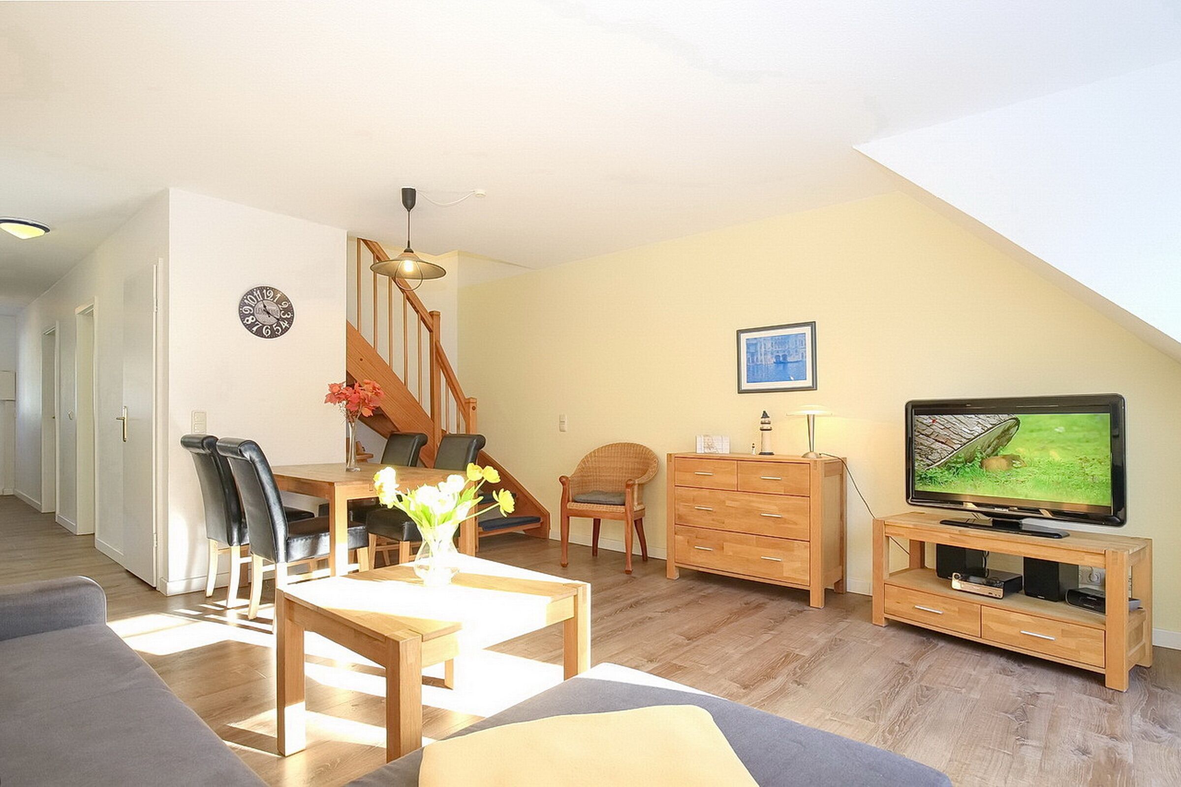 Un salon avec un escalier en bois, un coin repas, un téléviseur posé sur un meuble en bois et une horloge au mur.