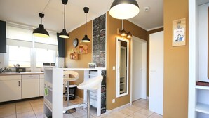 Une cuisine avec des armoires blanches, un plan de travail blanc et un tabouret de bar blanc. On y trouve des suspensions noires, un mur noir ardoise avec des inscriptions sur le thème du café, et une porte avec un miroir.