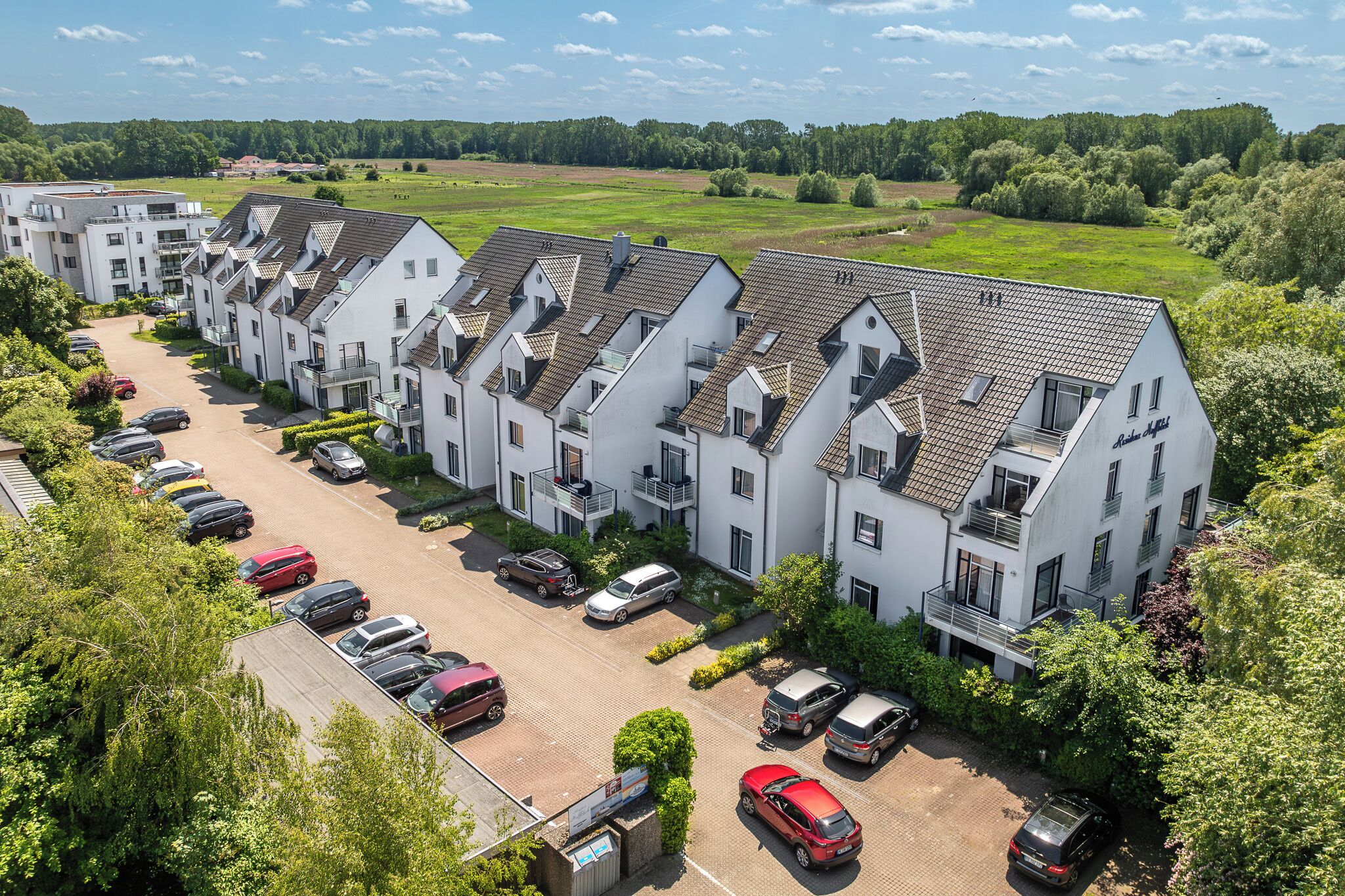 Een rij moderne, witte gebouwen met schuine daken, een parkeerplaats en een groen landschap op de achtergrond.