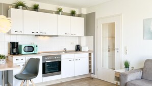 Una cocina funcional con armarios blancos, microondas, estufa y cafetera. En la sala de estar, hay una silla negra y un sofá gris.