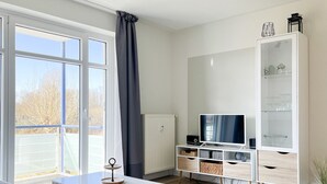 Una sala de estar moderna con un ventanal grande, un televisor de pantalla plana, un armario blanco con estantes y una mesita con un objeto decorativo.