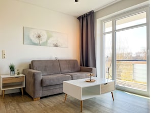 Moderni olohuone, jossa on harmaa sohva, valkoinen sivupöytä ja sohvapöytä. Suuresta ikkunasta avautuu näkymä ulos.