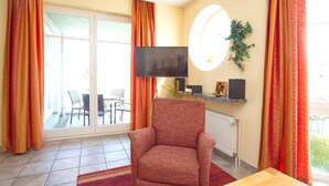 Un salon avec un fauteuil rouge, un téléviseur fixé au mur et une porte-fenêtre coulissante donnant sur une terrasse extérieure.