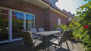 Een terras met een tafel en stoelen, een stenen huis en een tuin met een rode bloem.