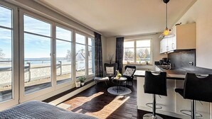 Ein modernes Wohnzimmer mit einem großen Fenster mit Blick auf einen Strand, ein Essbereich mit Barhocker und eine Küche mit Aussicht.