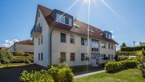 Un immeuble résidentiel à plusieurs étages, doté d’un toit en pente, d’un balcon au deuxième étage et d’un jardin bien entretenu.