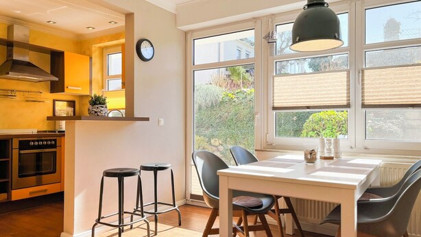 Une cuisine moderne dotée d’un four encastré, d’un coin repas avec une table et des chaises, et de grandes fenêtres équipées de stores.