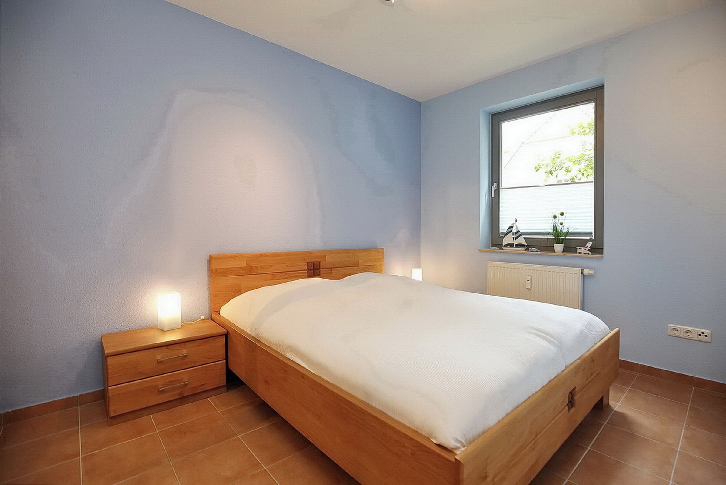 Un dormitorio con una cama de madera, una mesita de noche, una ventana con persianas y un radiador.