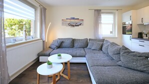 Een moderne woonkamer met een grijze modulaire bank, een witte salontafel met twee potplanten, een moderne hanglamp en uitzicht op een boot door een raam.