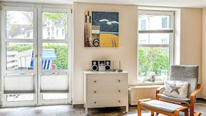 Une pièce avec une grande fenêtre, une commode blanche à tiroirs, une chaise avec un coussin et un tableau accroché au mur.