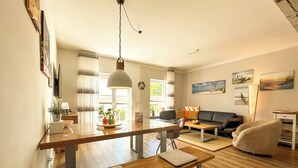 Een moderne woonkamer met een houten eettafel, een bank en een salontafel. Op de voorgrond staat een bankje en er hangt een lamp aan het plafond.