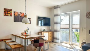 A modern kitchen with a wooden table, bench, and colorful chairs. There is a TV mounted on the wall and a window with curtains. - Landidyll Wohnung 02 in Elmenhorst (Elmenhorst)