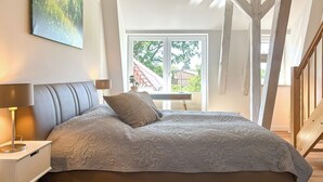 A bedroom with a large bed, a nightstand, a lamp, a window with a view of greenery, and a ceiling light. - Landidyll Wohnung 02 in Elmenhorst (Elmenhorst)