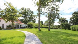 A paved pathway leading to a house with a red roof, surrounded by lush green grass and trees. - Landidyll Wohnung 03 in Elmenhorst (Elmenhorst)