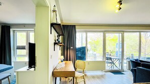 Een kamer met een houten bureau, een stoel, een bed en een bank. Er is een raam met gordijnen, een lamp en uitzicht op het omliggende landschap.