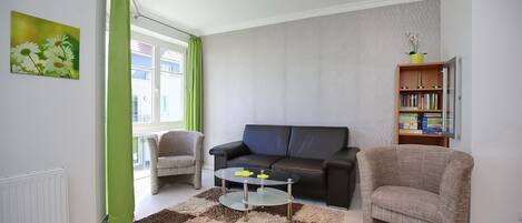 Una sala de estar con sofá, sillones, una mesa de centro, una estantería y un ventanal con cortinas.