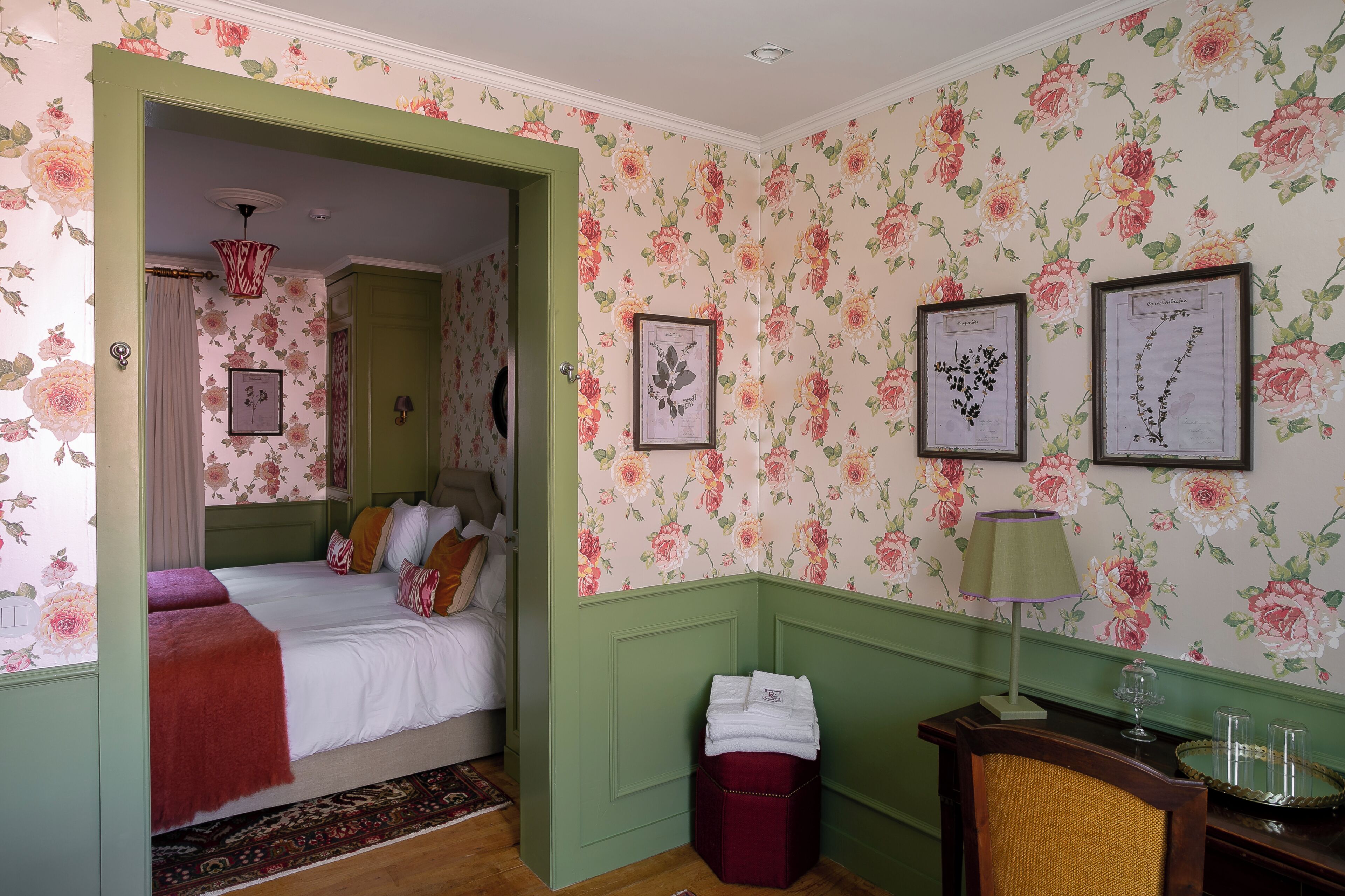 A bedroom with floral wallpaper, a bed with pillows, a nightstand, a lamp, and a framed botanical print on the wall.