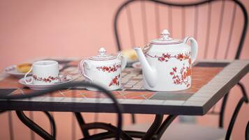 Ein Tisch mit Teekanne und Kaffeegedeck auf einer gefliesten Tischdecke, ein Stuhl und ein Teller mit einer Brotscheibe.