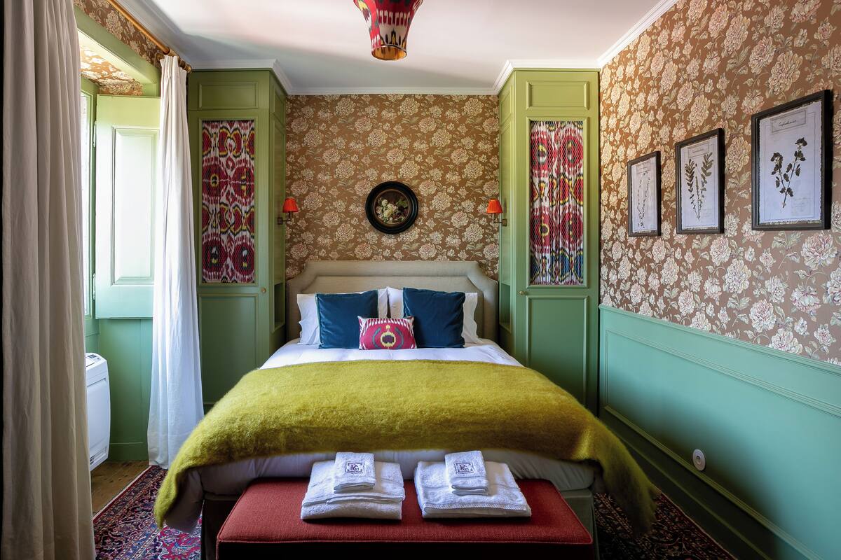 A neatly arranged bedroom with a bed, pillows, a bedside table with towels, and a wall-mounted clock.